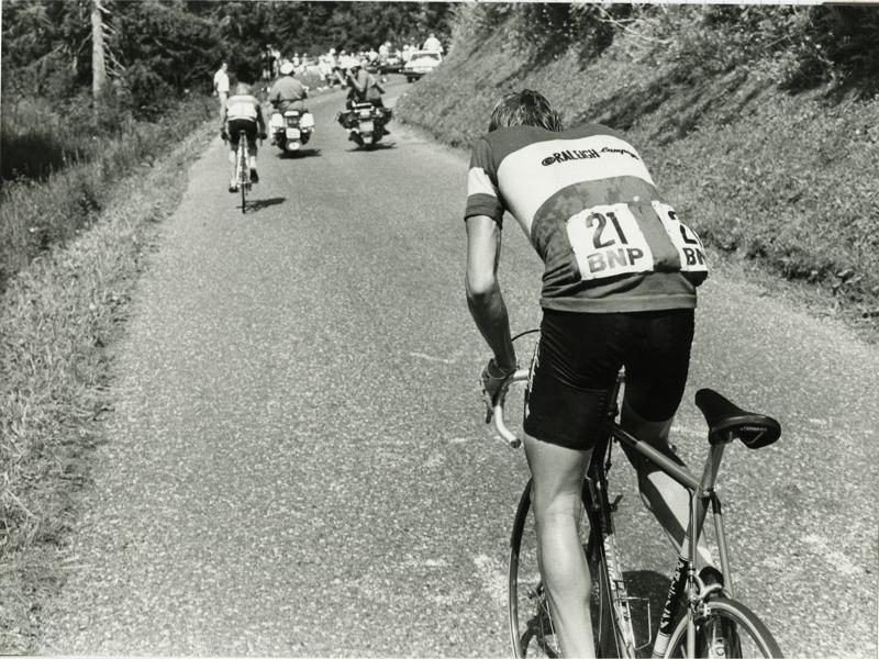 Archival photograph of a lone road cyclist, seen from behind, on a long open road